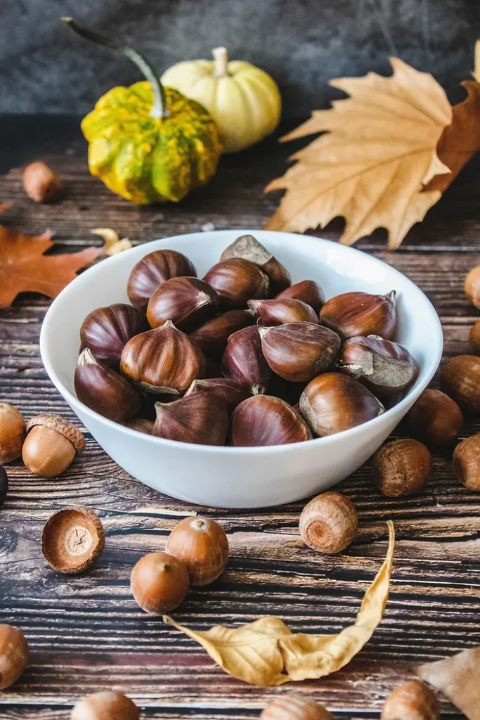 Bowl of chestnuts with hazelnuts, a gourd and autumn leaves on a wooden table